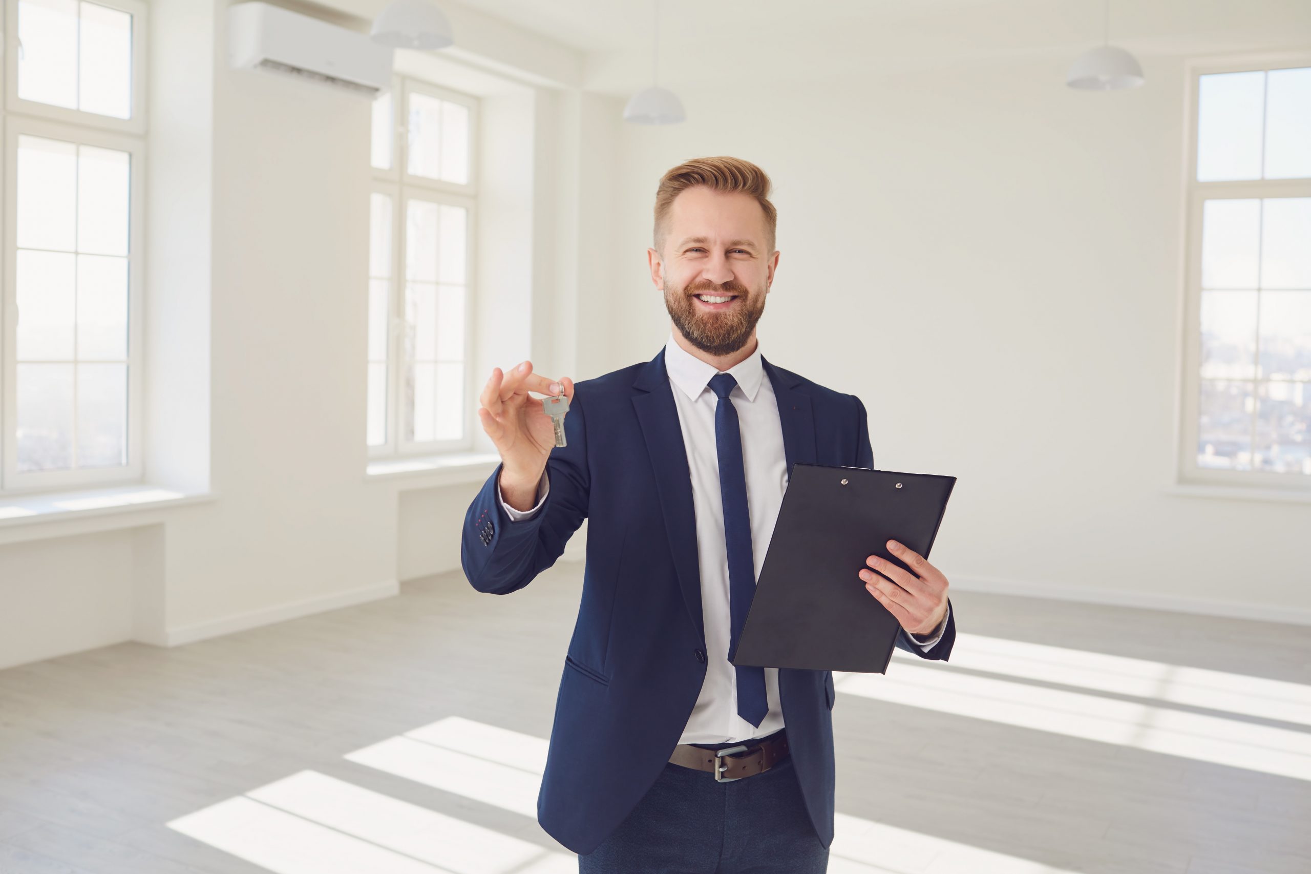Successful salesman in trendy suit holding clipboard and key smiling at camera in fresh new apartment